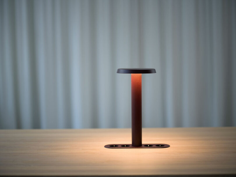 Una moderna lámpara de mesa rectangular con una base marrón y un poste vertical se asienta sobre una superficie de madera, iluminando suavemente el área. El fondo es una simple cortina de luz.