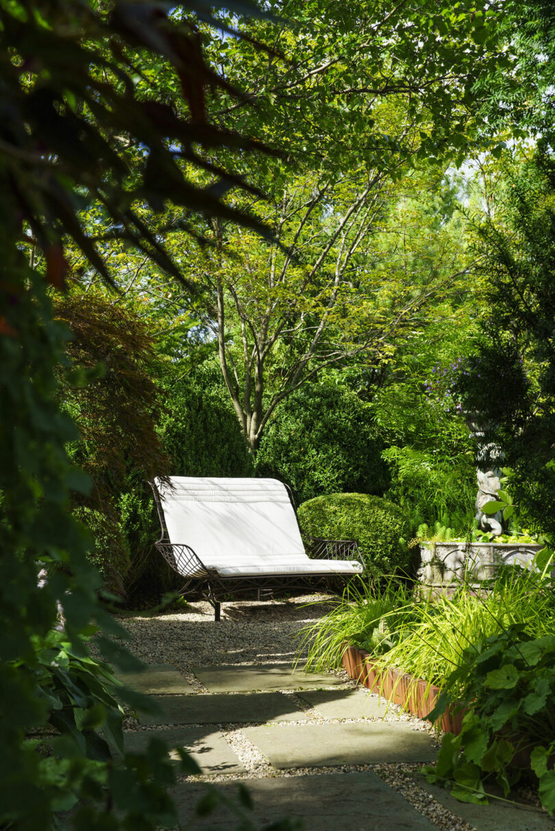 Jardín soleado con bancos blancos rodeados de frondosos árboles, arbustos y plantas verdes, con escalones de piedra y grava en el suelo.