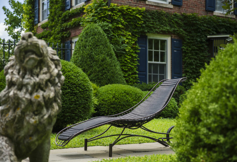 Un sillón de metal negro se encuentra en una terraza de piedra, rodeado de arbustos y plantas bien cuidados, estatuas de leones de piedra y una casa de ladrillo cubierta de hiedra.