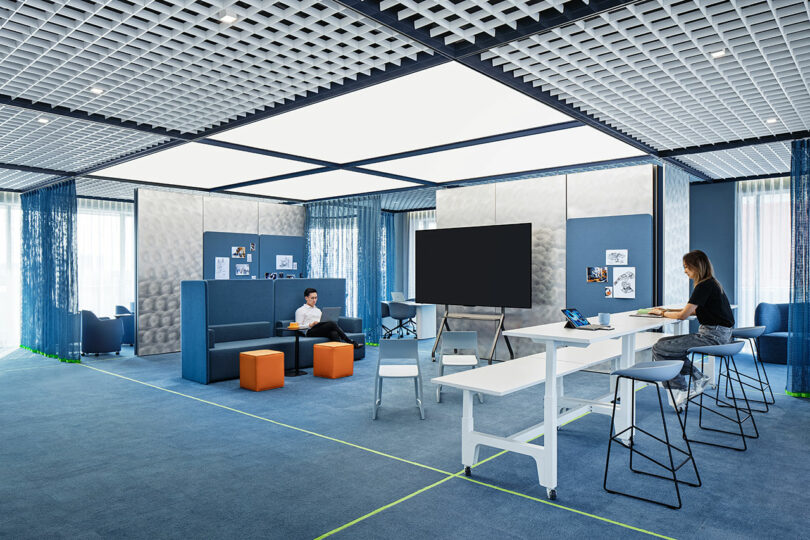 Espacio de oficina moderno con alfombra azul, sillas modulares, mesa, taburetes y dos personas trabajando con portátiles. En el centro de la sala se ven una gran pantalla y una pizarra.