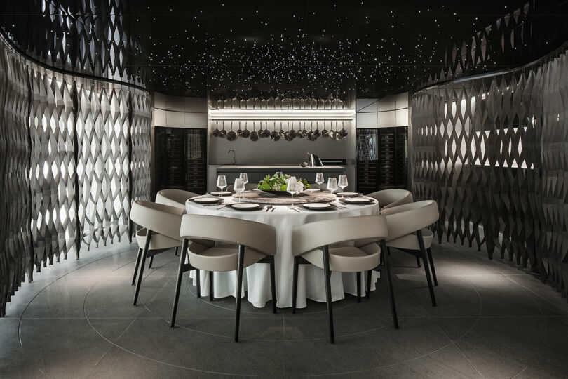Una mesa de comedor redonda para seis personas con modernas sillas blancas en una habitación oscura y elegante con paredes con dibujos oscuros y pequeñas luces en forma de estrella en el techo.