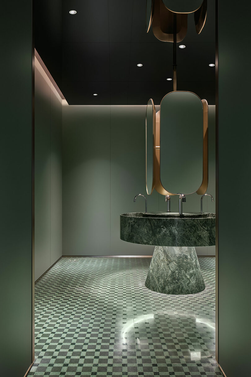 El baño moderno cuenta con un lavabo con pedestal de mármol verde, un espejo geométrico, baldosas de cuadros verdes y una iluminación minimalista.