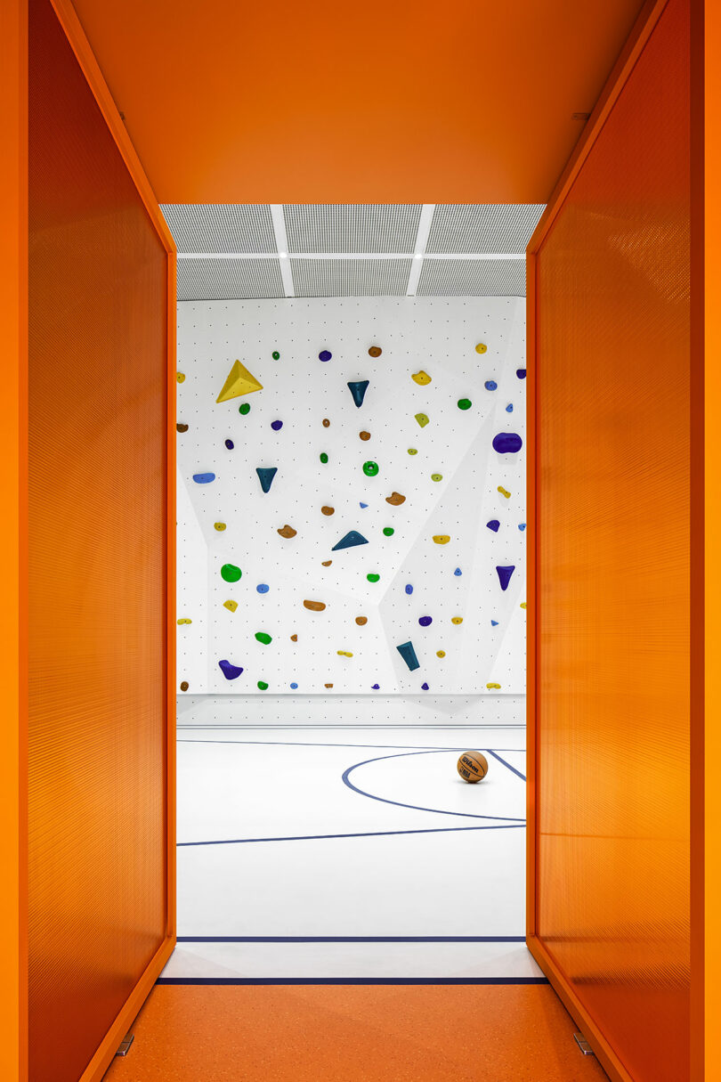 El gimnasio se puede ver a través de las puertas dobles de color naranja, que cuenta con un muro de escalada blanco con gradas de colores y una pelota de baloncesto sobre una cancha blanca con líneas azules.