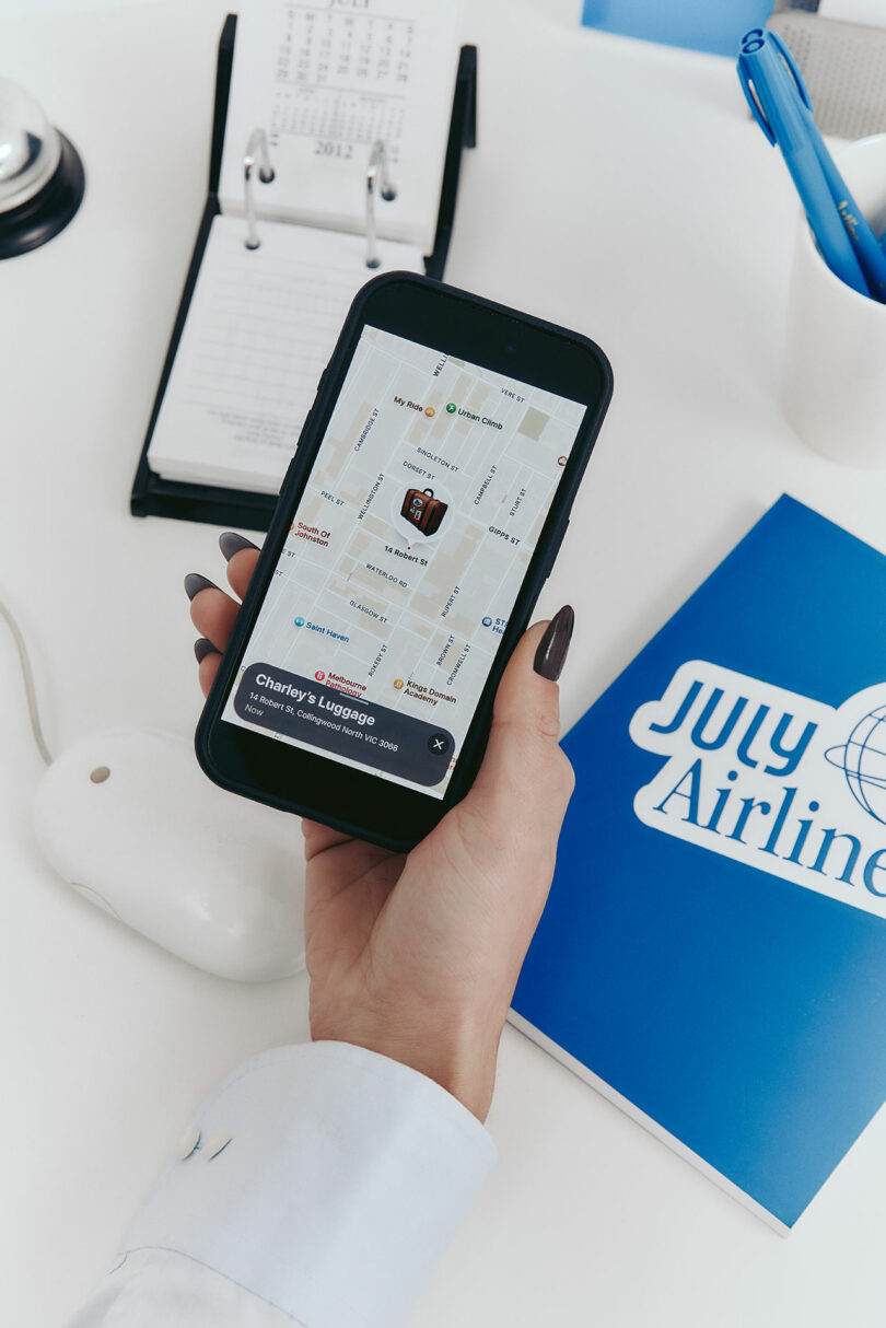 Una persona sostiene un teléfono inteligente que muestra un mapa que rastrea la ubicación del equipaje; un calendario, un mouse y carpetas de July Airlines sobre una mesa blanca.