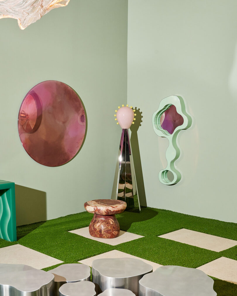 La moderna habitación presenta una decoración geométrica que incluye espejos redondos de color rosa, arte abstracto en las paredes de color verde, taburetes de color rosa y marrón, superficies metálicas y un piso de césped artificial con baldosas blancas.