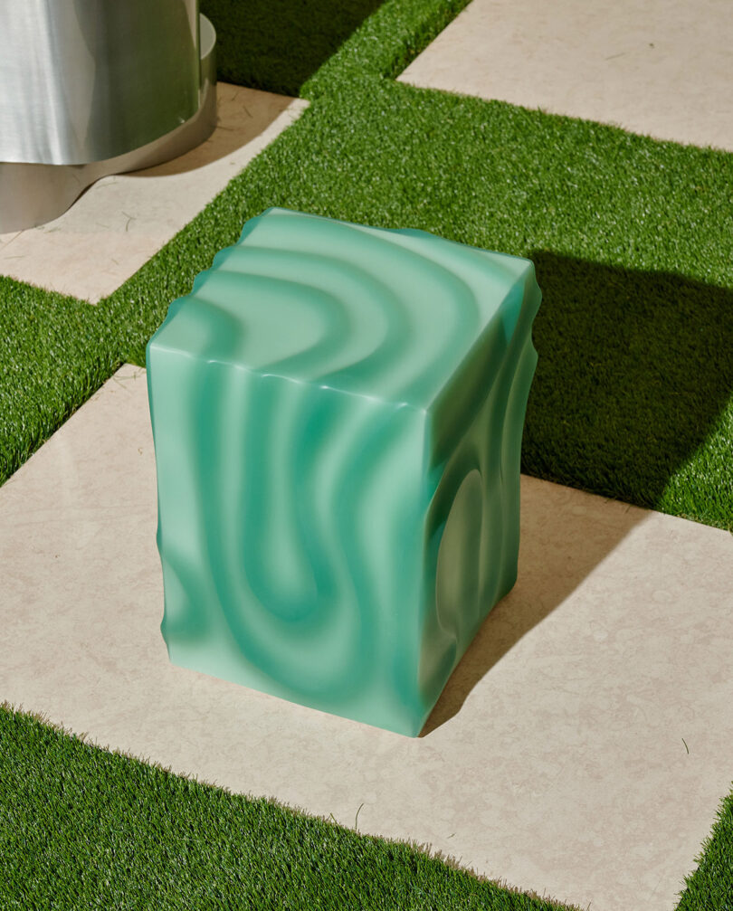 Un objeto verde con forma de cubo con estampado de ondas se encuentra en una terraza de azulejos bordeada de césped artificial.