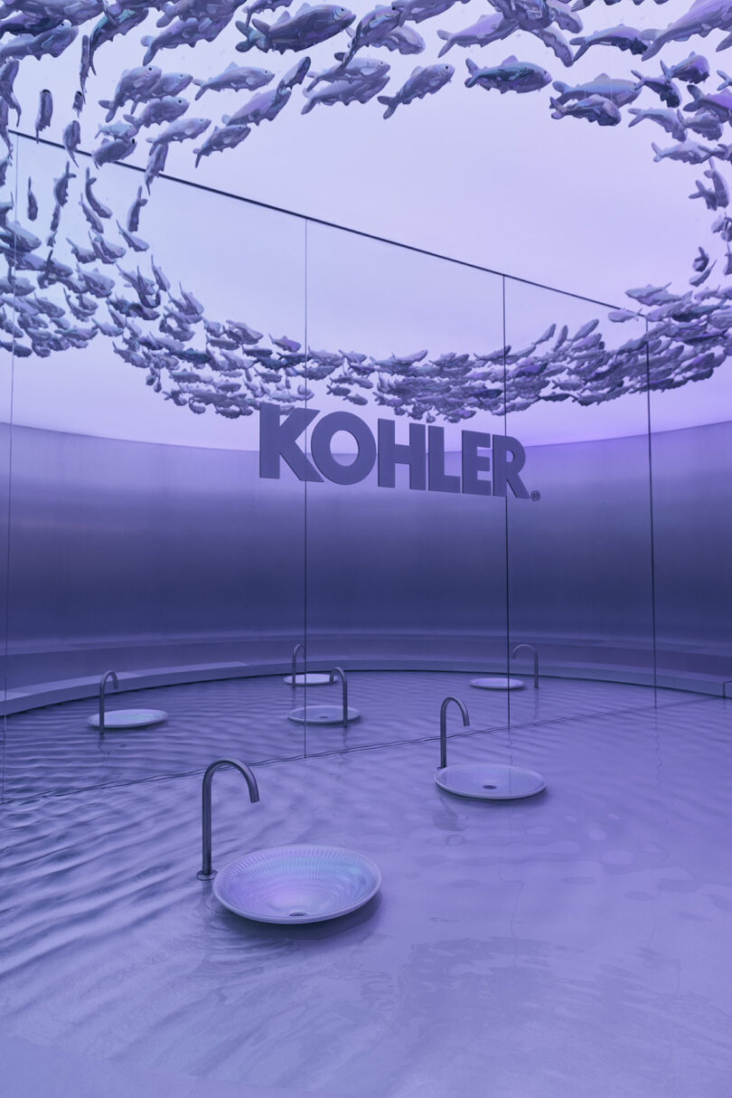 Sala de exposición moderna con cuatro lavabos redondos, grifos metálicos y paredes reflectantes "Kohler" El logotipo de la marca y la escultura en forma de pez que cuelga del techo están bañados por una luz violeta.