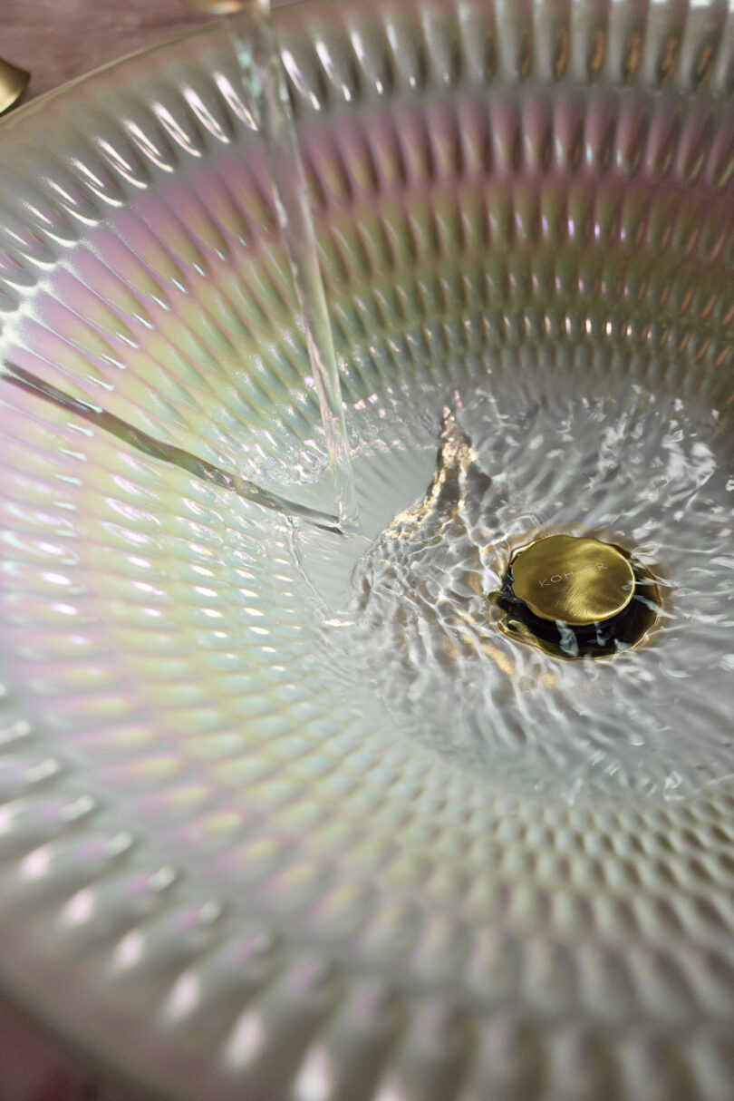 El agua fluye hacia un fregadero iridiscente texturizado con una tapa de drenaje dorada, creando ondas en la superficie.