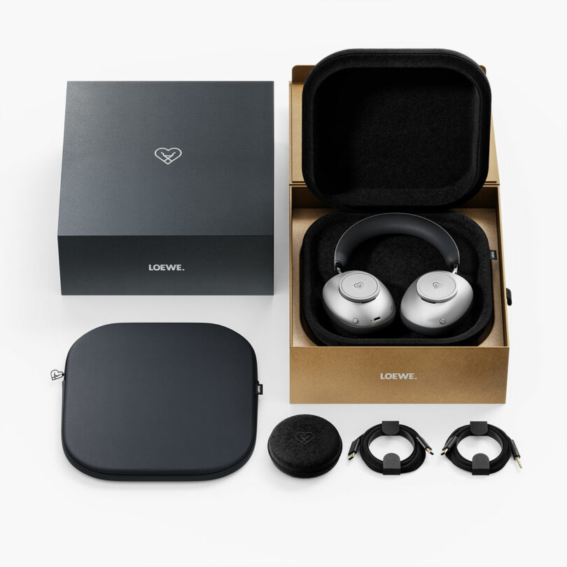 Un par de auriculares supraaurales plateados vienen en una caja abierta, que contiene una caja negra, dos cables, una pequeña bolsa redonda y un embalaje adicional, cuidadosamente dispuestos sobre una superficie blanca.