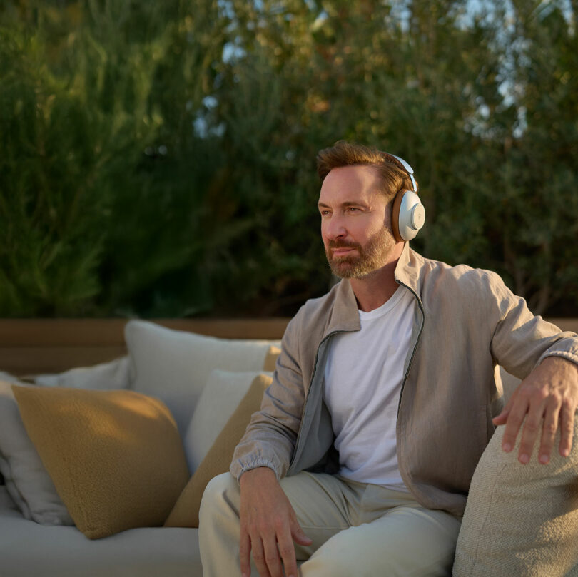 Un hombre con auriculares inalámbricos está sentado en un sofá al aire libre con cojines beige, rodeado de plantas verdes.