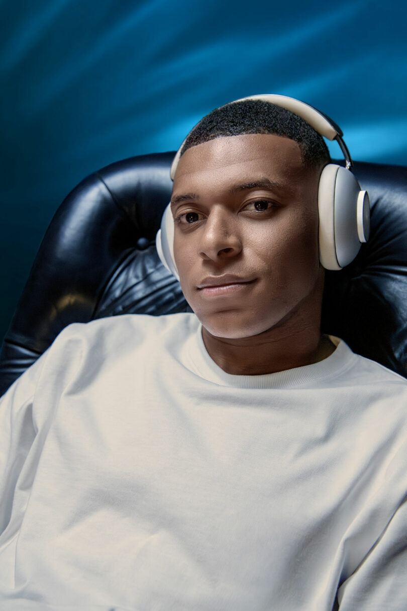 Un hombre con una camisa blanca y grandes auriculares blancos se sienta en una silla de cuero negro y mira la cámara sobre un fondo azul.