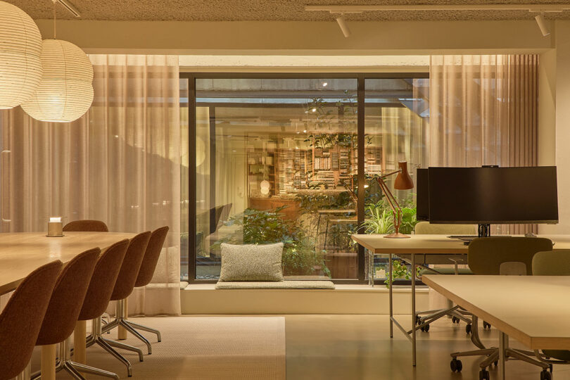 Espacio de oficina moderno con mesa de conferencias, sillas, monitor de computadora en el escritorio, iluminación tenue y grandes ventanales con cortinas transparentes que dan a un patio verde.
