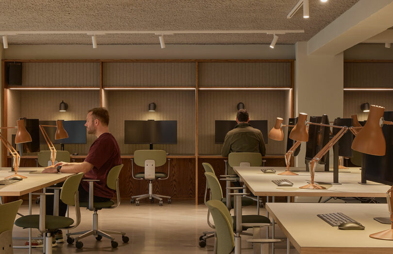 Espacio de oficina moderno con escritorio de madera, silla verde y lámpara marrón; dos personas trabajando por separado frente a la computadora en una habitación bien iluminada.
