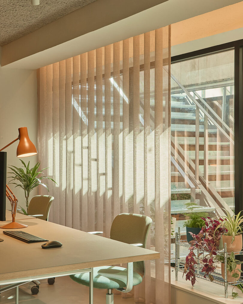 Oficina moderna con dos sillas verdes, un escritorio, una computadora, una lámpara, plantas en macetas y persianas verticales que cubren parcialmente una gran ventana con escaleras visibles desde el exterior.