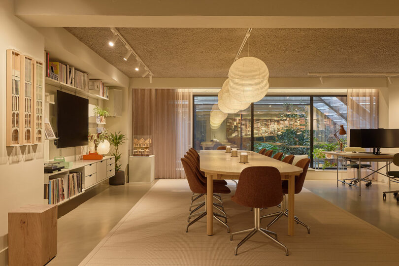 Espacio de oficina moderno con una larga mesa de madera, sillas marrones, faroles de papel colgantes, estanterías, TV, plantas de interior, grandes ventanales y vistas al verde jardín exterior.