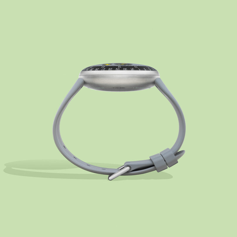 A side view of a modern TYPE 3 wristwatch with a grey strap and a dark round face, displayed against a light green background.