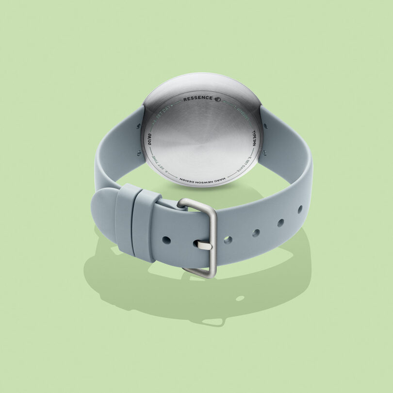 A TYPE 3 wristwatch with a light gray strap is displayed face-down on a light green background, showing the metal case back and buckle.