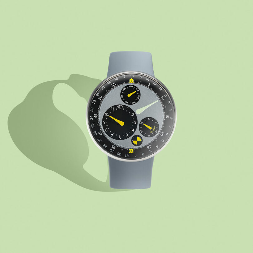 A modern TYPE 3 wristwatch with a gray strap and a black dial featuring multiple subdials and yellow details, displayed on a light green background.