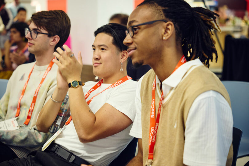 Three people wearing NeoCon conference lanyards sit in a row; one is clapping while the others watch attentively.