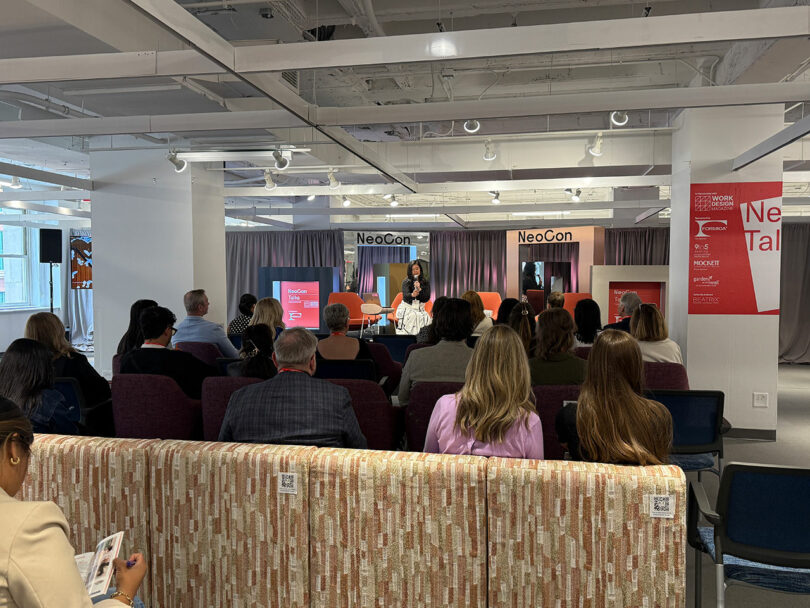 Un grupo de personas se sienta frente a un escenario donde un orador habla en NeoCon, un evento que se lleva a cabo en un espacio de conferencias moderno y lleno de luz.