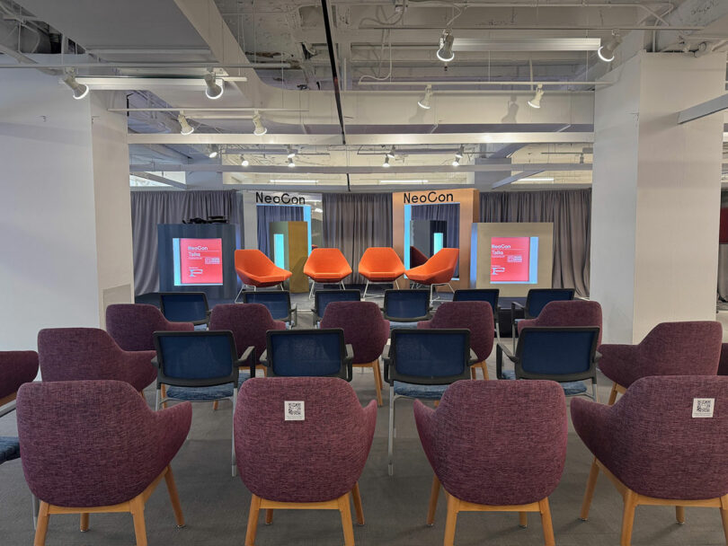 Una moderna sala de conferencias con una hilera de sillas moradas frente a un escenario con cinco sillas naranjas, dos pantallas y un letrero neoconservador en lo alto.