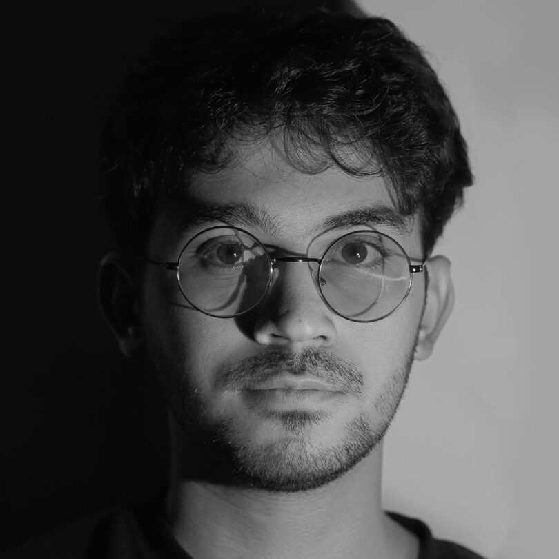 Un joven de pelo corto, gafas y barba mira directamente a la cámara. La luz creó un fuerte contraste de luces y sombras en su rostro.