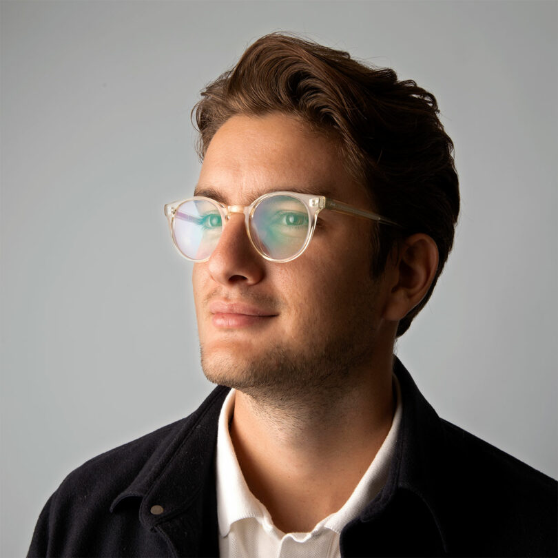 Un joven de cabello castaño y gafas, vestido con camisa blanca y chaqueta oscura, mira ligeramente hacia arriba sobre un fondo liso.