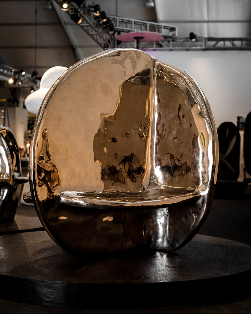 Un banco escultórico de metal pulido con una superficie reflectante redondeada exhibido en un espacio de exposición interior.