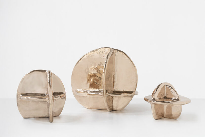 Tres esculturas abstractas de bronce metálico, de diferentes tamaños, con planos curvos y rectilíneos que se cruzan, expuestas sobre una superficie de color blanco puro sobre un fondo blanco.