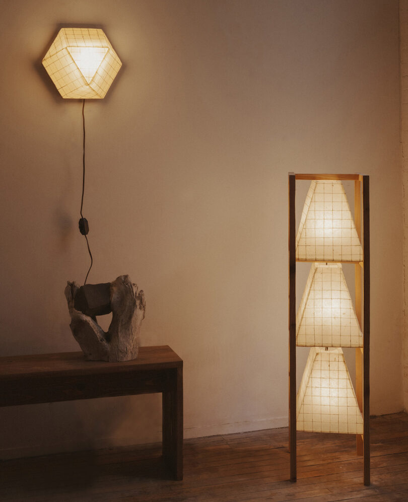 La sala minimalista, amueblada con taburetes de madera y esculturas de madera flotante, está iluminada por un aplique de pared hexagonal y una lámpara de pie con tres pantallas en forma de pirámide.