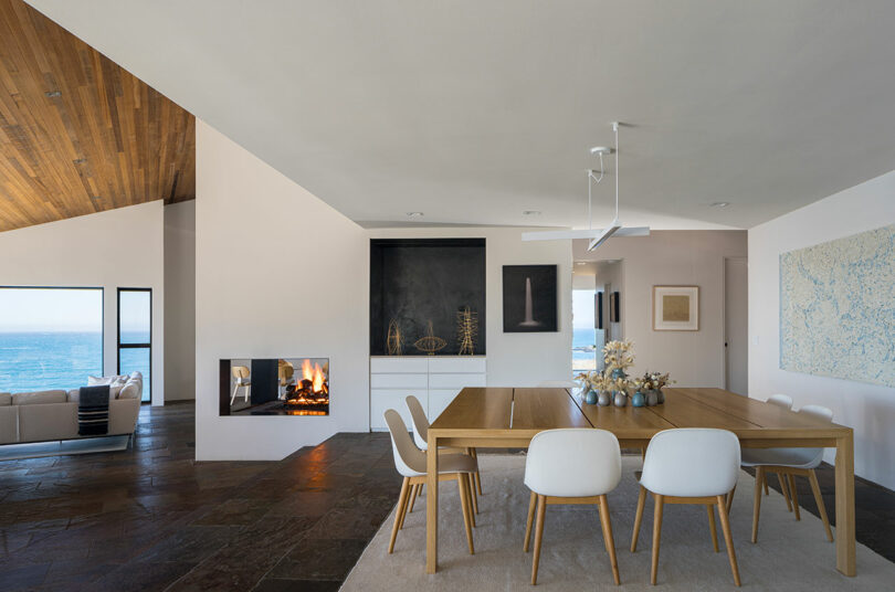 Comedor moderno de planta abierta con mesa de madera, sillas blancas, chimenea incorporada y vistas al mar a través de grandes ventanales. Decoración minimalista con arte abstracto en las paredes.