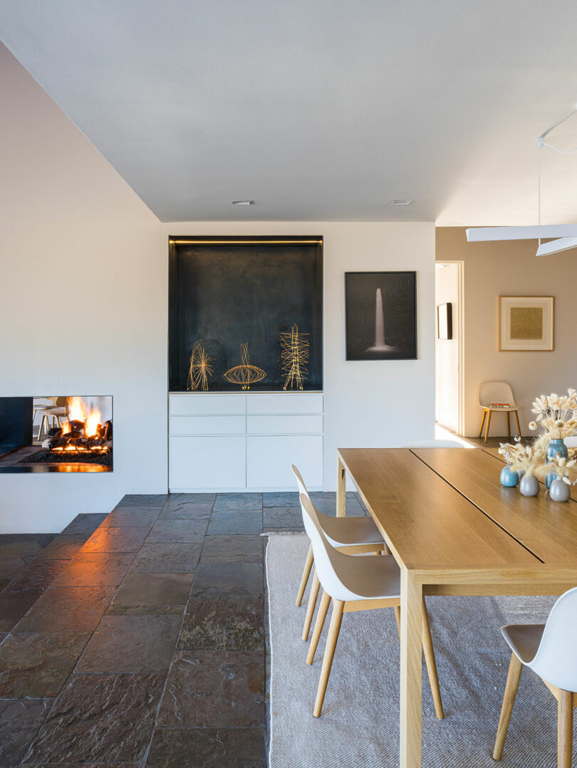 Comedor moderno con mesa de madera, sillas blancas, adornos en los estantes, suelo de baldosas de piedra y chimenea encendida en el lado izquierdo.