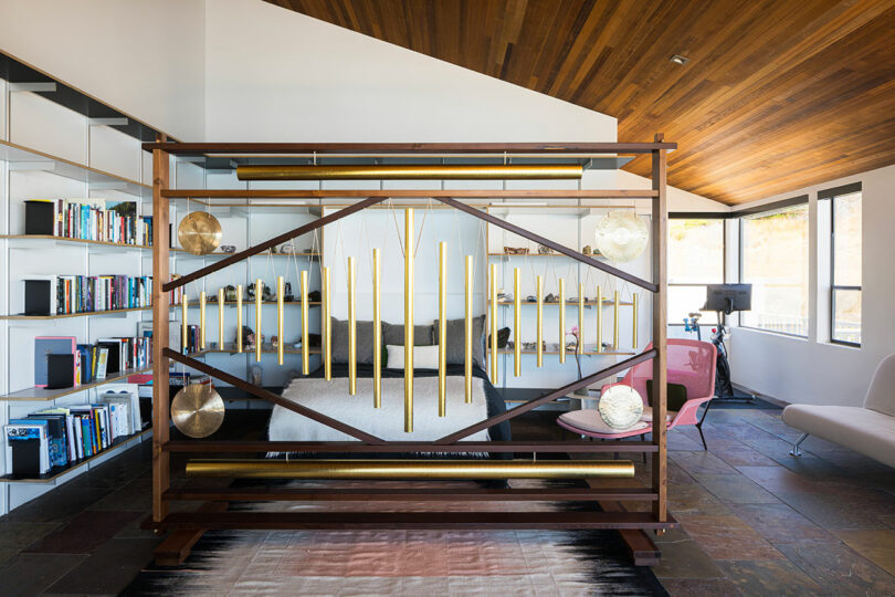 Un dormitorio moderno con una cama separada por un tabique decorativo de madera y latón con estanterías a la izquierda y asientos junto a una gran ventana a la derecha.