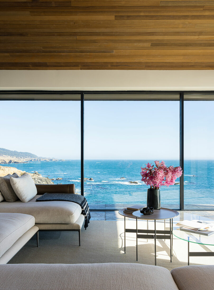 Sala de estar moderna con sofá seccional beige, mesa de centro de vidrio decorada con flores rosas y grandes ventanales con vista al océano.