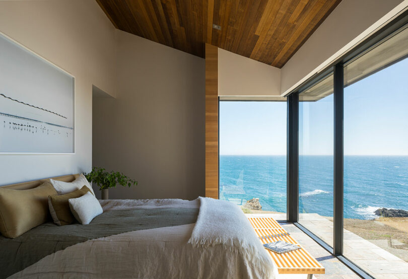 El moderno dormitorio cuenta con grandes ventanales con vistas al mar, una cama con ropa de cama de color neutro, techos de madera y una decoración minimalista.