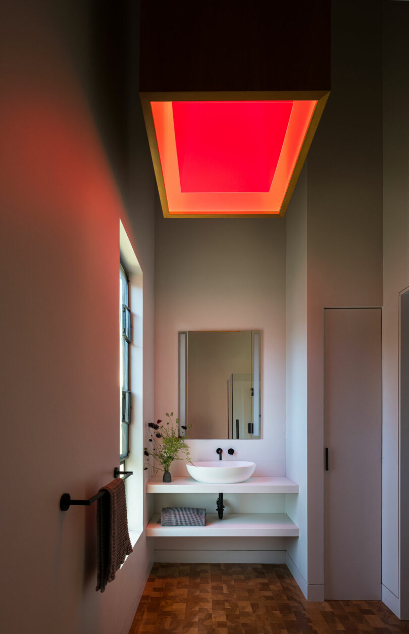 El moderno baño cuenta con un lavabo blanco flotante, un espejo rectangular y un gran panel de luz en el techo que brilla en rojo. Una toalla cuelga a la izquierda y la ventana deja entrar luz natural.