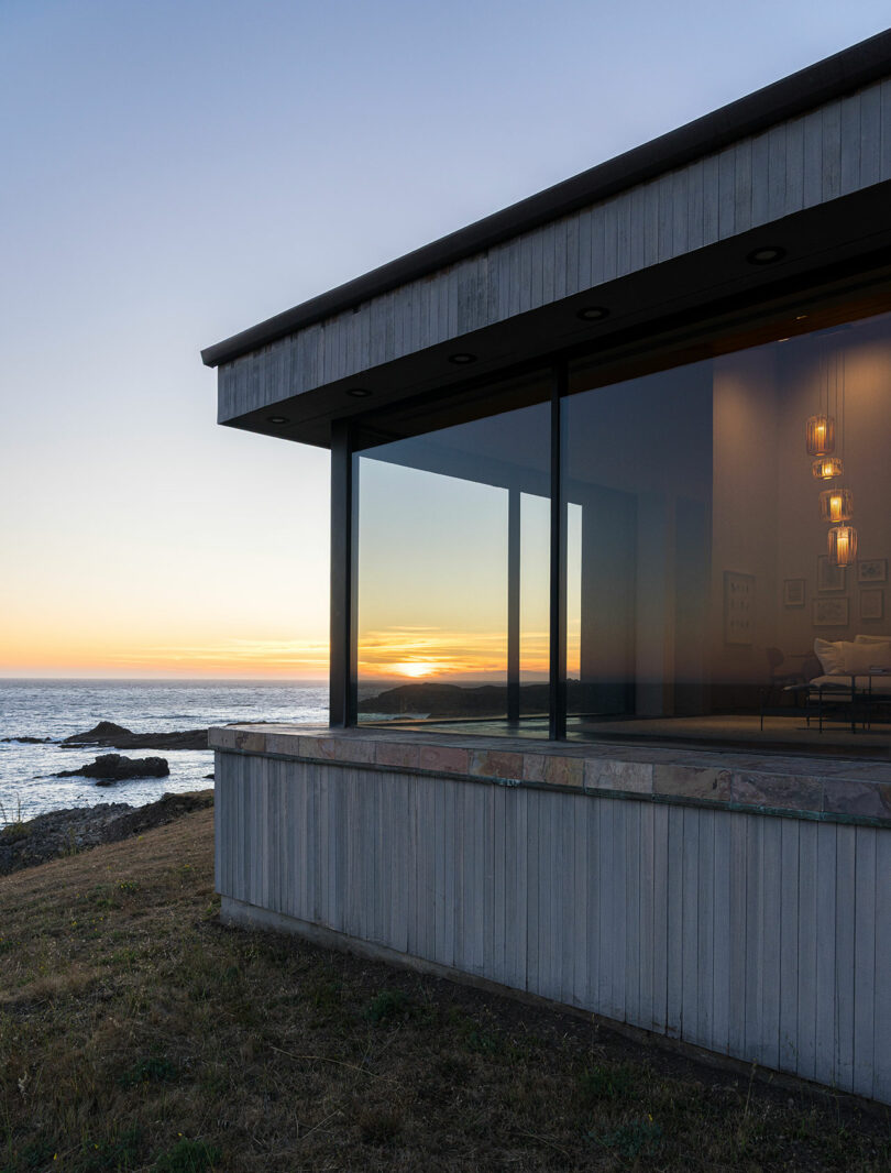 Casa moderna con grandes ventanales con vistas al mar al atardecer con cálidas luces interiores y prados en primer plano.