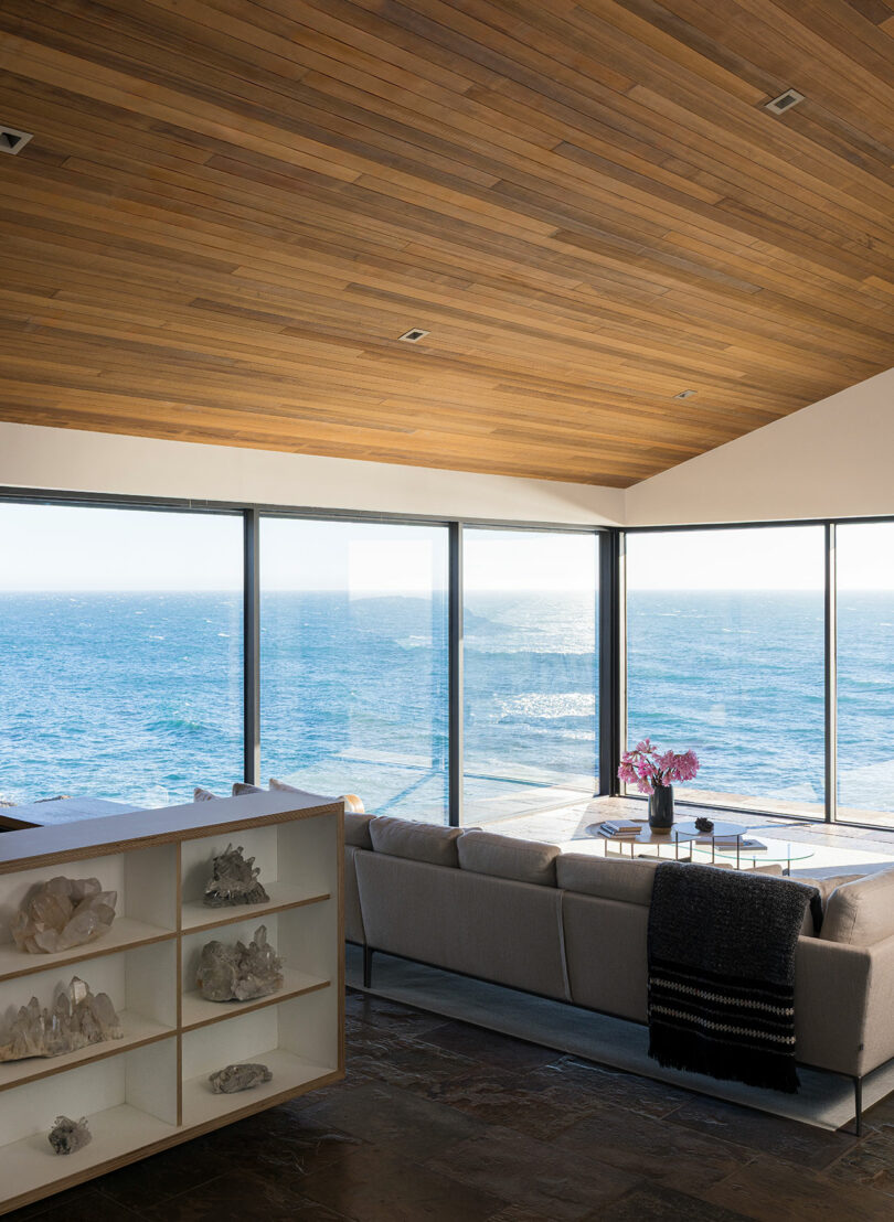 La moderna sala de estar cuenta con ventanales con vista al océano, un sofá gris claro, una mesa de café llena de flores y un expositor de cristal.