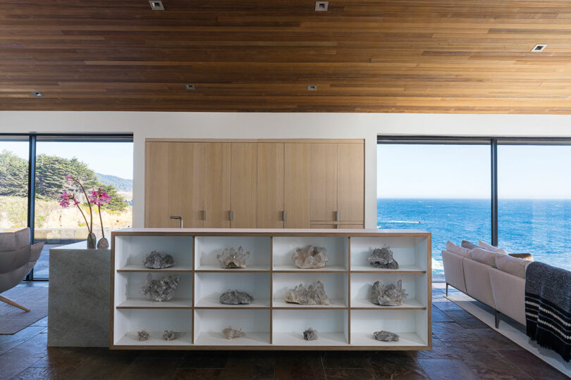 Salón moderno con techo de madera, estanterías abiertas con corales, grandes ventanales con vistas al mar y mobiliario minimalista.