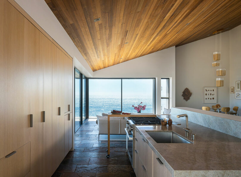 La moderna cocina tiene suelos de piedra, techos con paneles de madera y grandes ventanales con vistas al mar. Cerca de la ventana se ven una pequeña zona para sentarse y una mesa de comedor.