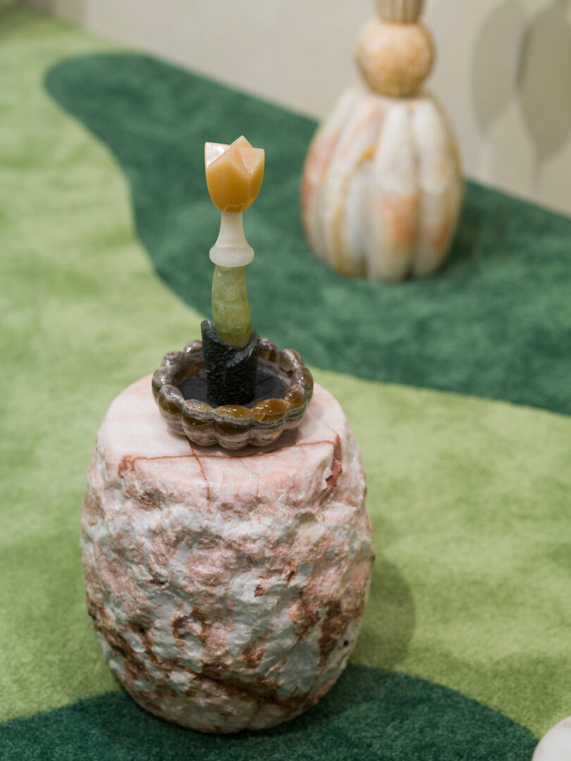 Una pequeña escultura de Sten Studio hecha de piedra y minerales, con forma de flor, se asienta sobre una base de piedra texturizada, cubierta con una alfombra verde, y otra escultura al fondo.