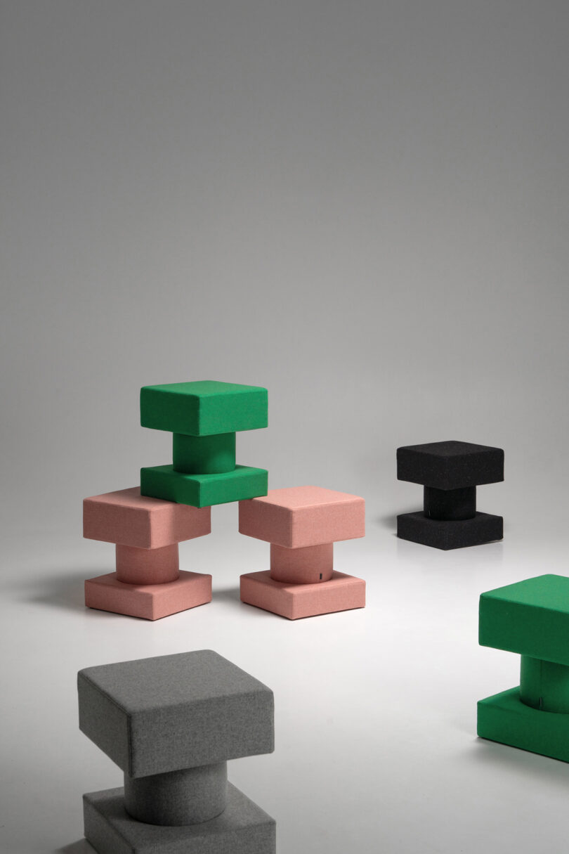 Geometric stools in green, pink, black, and gray are arranged on a plain, light-colored surface against a neutral background.