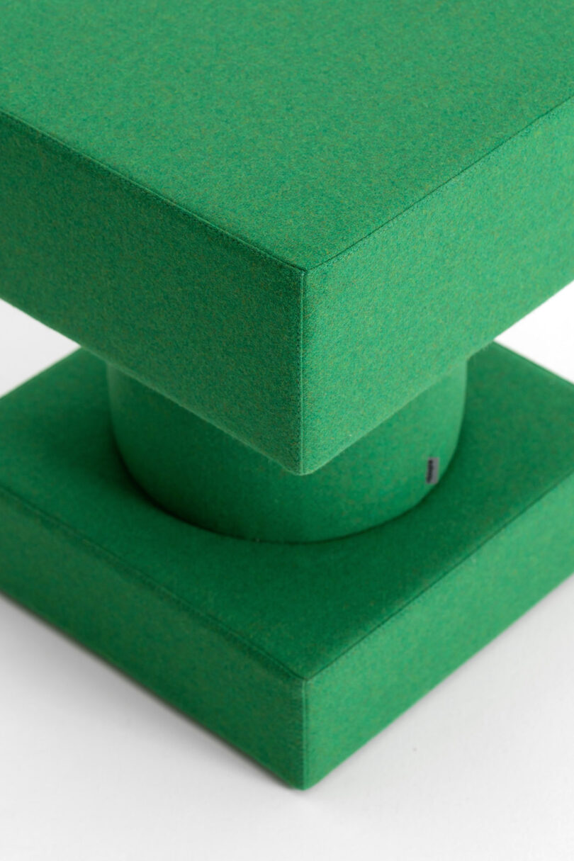 Green geometric upholstered object with a square top, cylindrical middle, and square base, placed on a white surface.