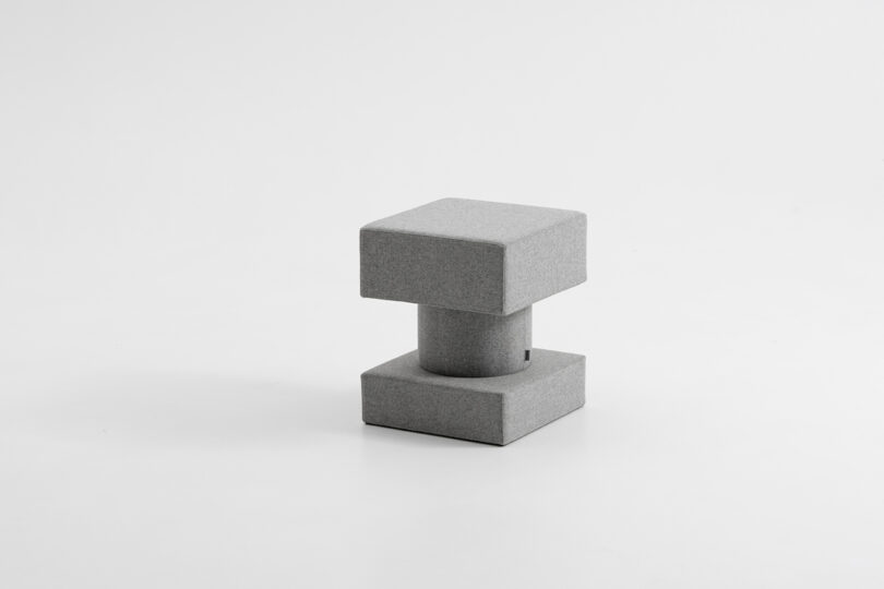 A small, modern, gray upholstered stool with a square seat, cylindrical middle, and square base on a plain white background.