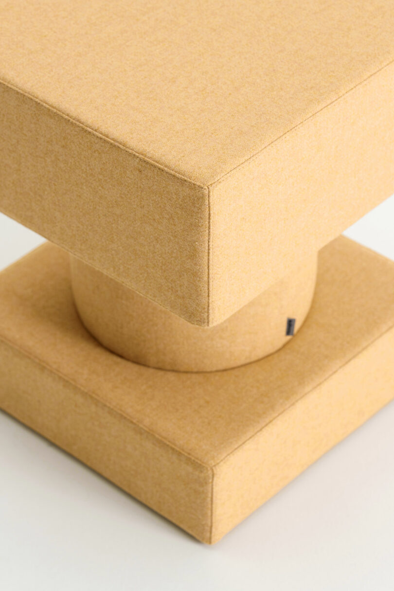 A geometric, mustard-yellow upholstered stool with a square top and base connected by a cylindrical center.