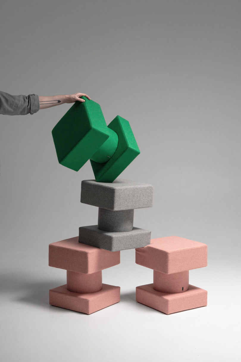 A person’s hand places two green foam blocks on top of a stack of gray and pink square foam blocks arranged in a balanced structure against a plain background.