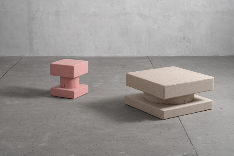 Two geometric sculptural stools or tables, one pink and one beige, placed on a gray concrete floor against a textured gray wall.
