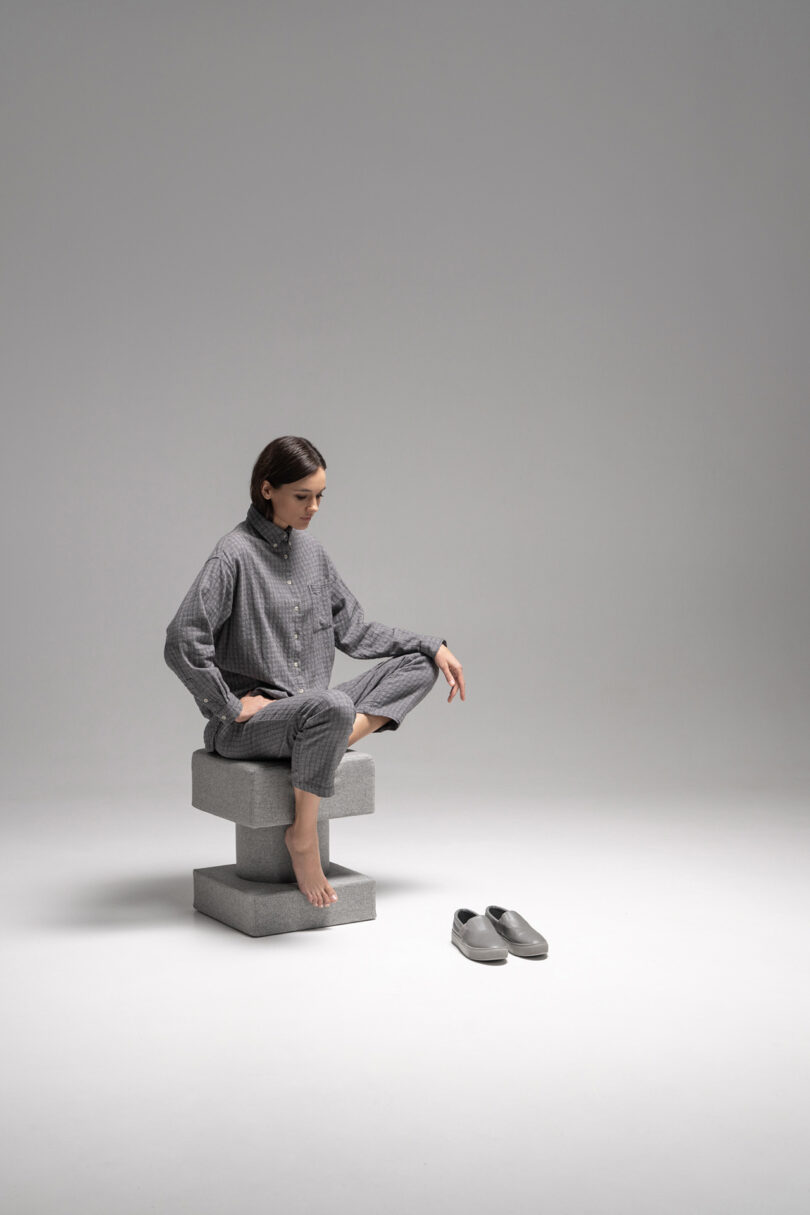A woman in gray pajamas sits cross-legged on stacked gray blocks, with her bare feet visible and gray slippers placed on the floor in front of her.