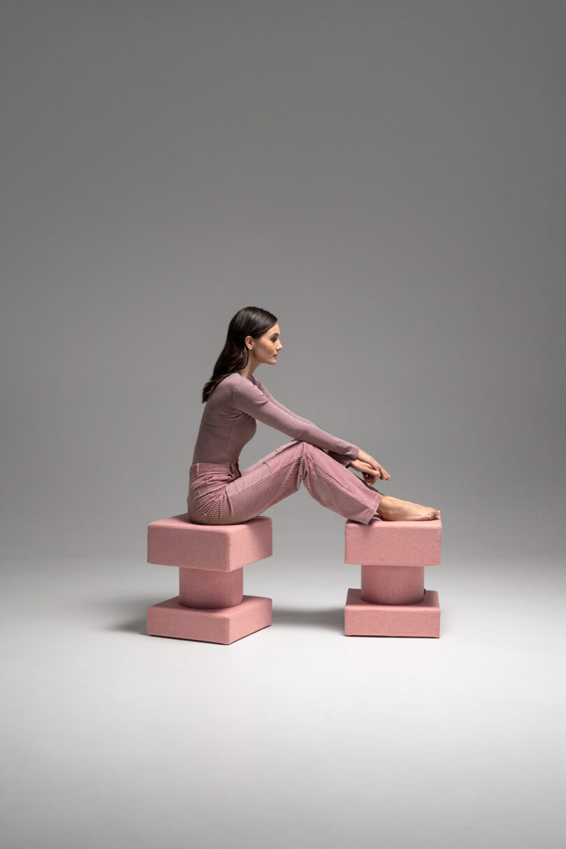 A woman in pink clothing sits sideways with her knees up on two geometric pink blocks in a minimalist studio setting.