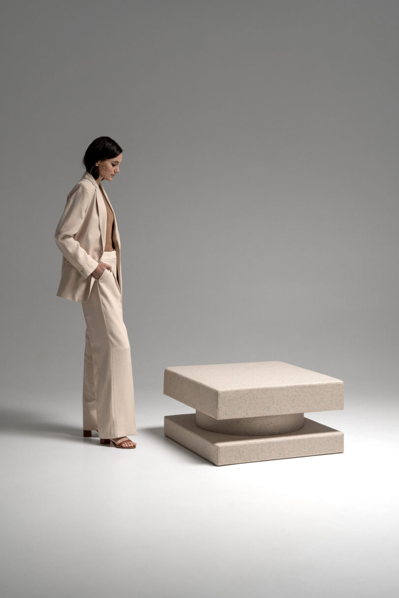 A woman in a beige suit stands in a minimalist studio, looking at a modern, square, layered beige table on a white floor with a neutral background.